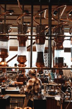 A barista working amidst large coffee machines and copper pipes in a stylish brewery-style cafe.