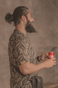 Bearded man with man bun uses smartphone and holds soda can in relaxed indoor setting.