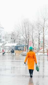 A woman in an orange coat walks on a snowy, wet city street, capturing a winter day vibe.