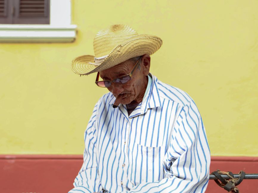 An elderly man in a straw hat and striped shirt, smoking outdoors against a yellow wall.
