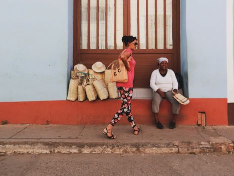 A woman walks along a vibrant street with a vendor selling woven baskets in the background.