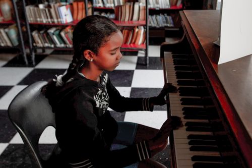 Free A young girl focused on playing a piano in a library, blending music with literature. Stock Photo