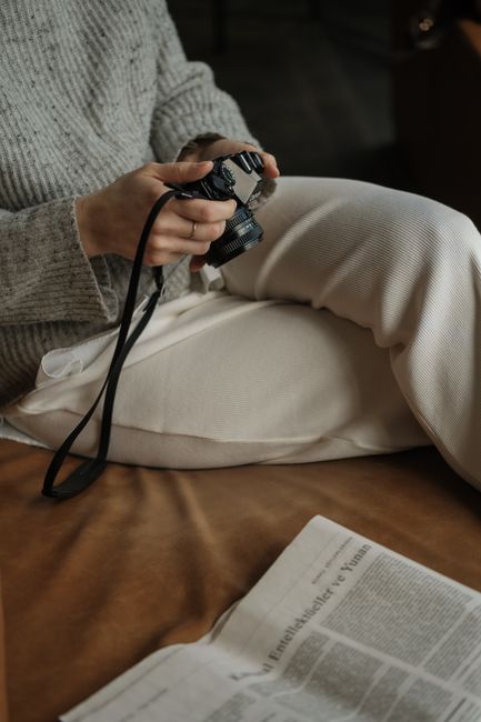 A woman sitting on a couch holding a camera with a nearby newspaper, creating a cozy indoor scene.
