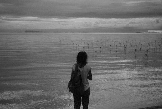 Rear view of a woman with a backpack gazing at the serene ocean. Moody black and white.
