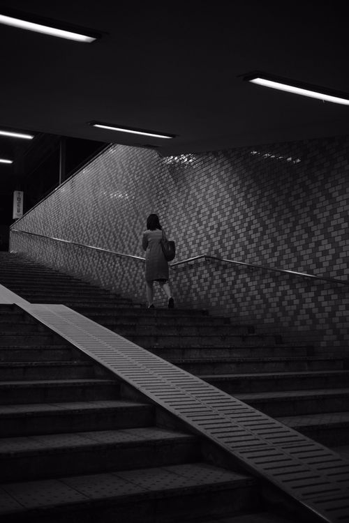 Free A solitary woman walks up subway stairs in a dark, monochrome setting. Stock Photo