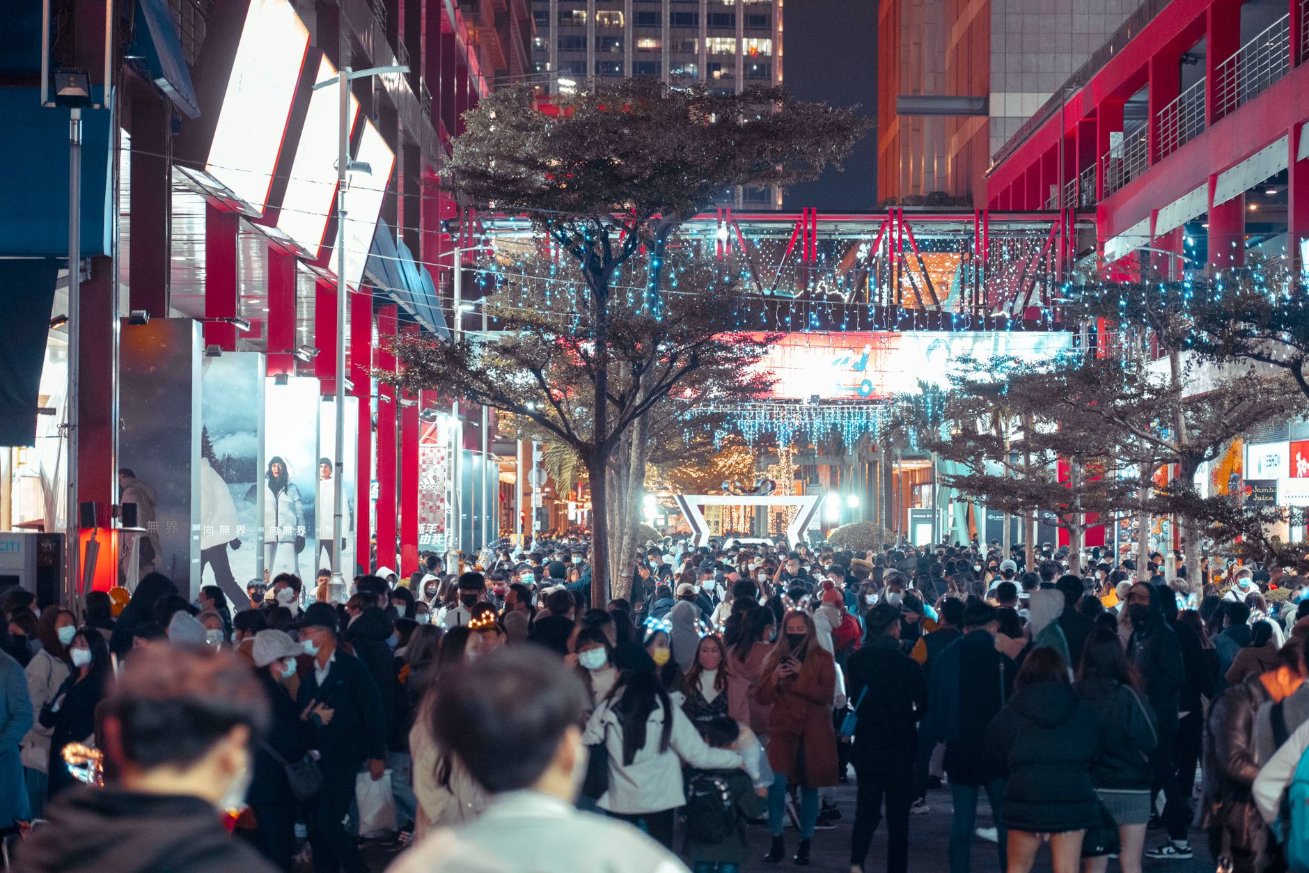 A bustling urban street filled with people at night, illuminated by city lights and decorations.