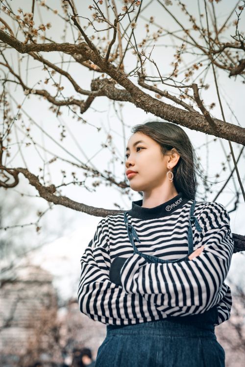 Free Young woman standing with arms crossed under blooming tree branches in spring. Stock Photo