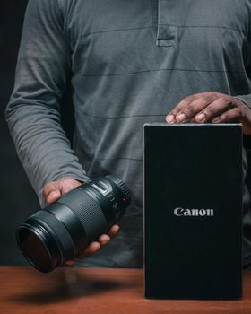 Detailed photo of hands holding a camera lens beside its Canon box on a wooden surface.