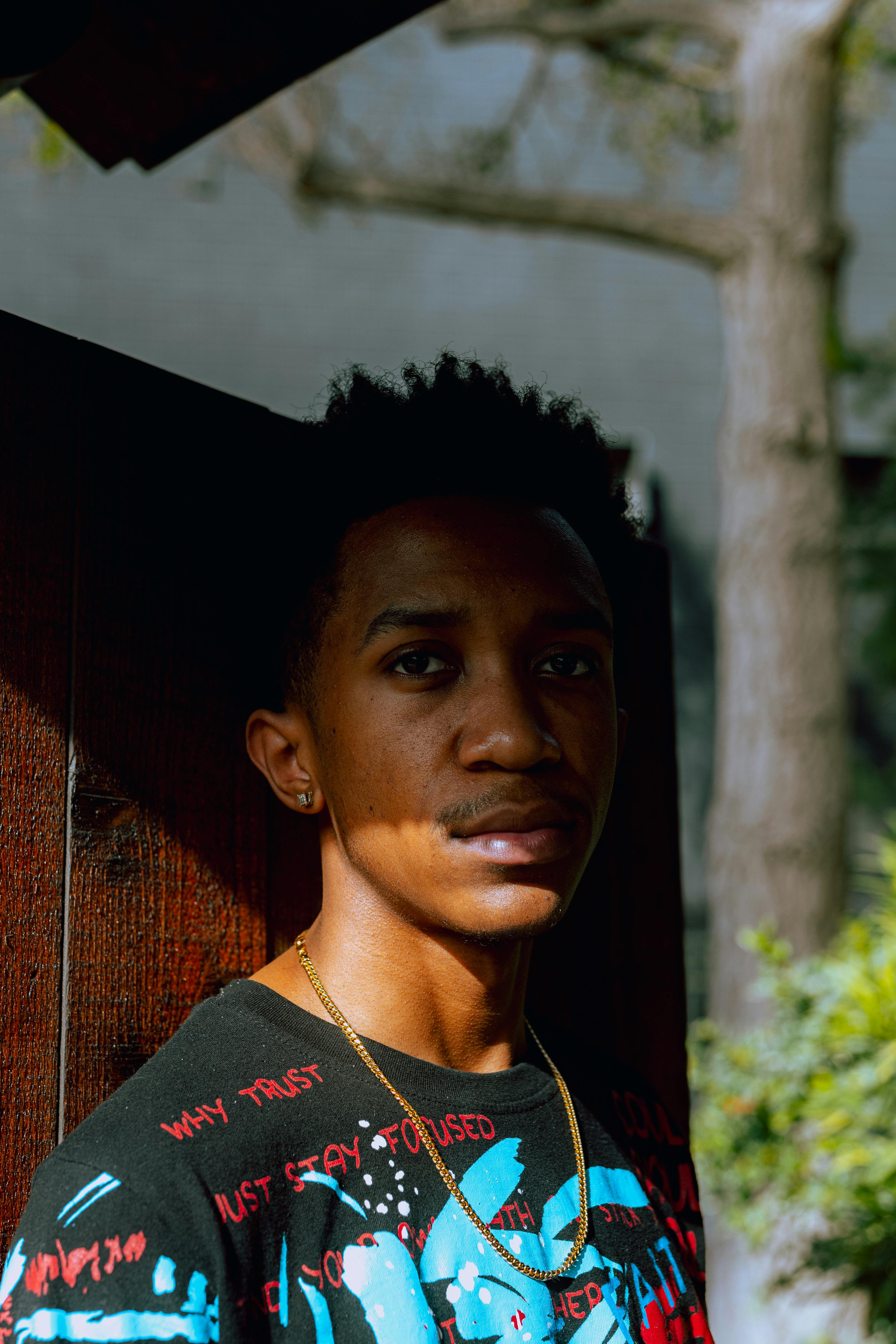 A young man stands against a wooden background with sunlight casting shadows across his face.