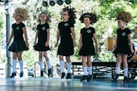 一位年轻女孩头戴假发，身着传统服装，在户外舞台上表演爱尔兰舞蹈