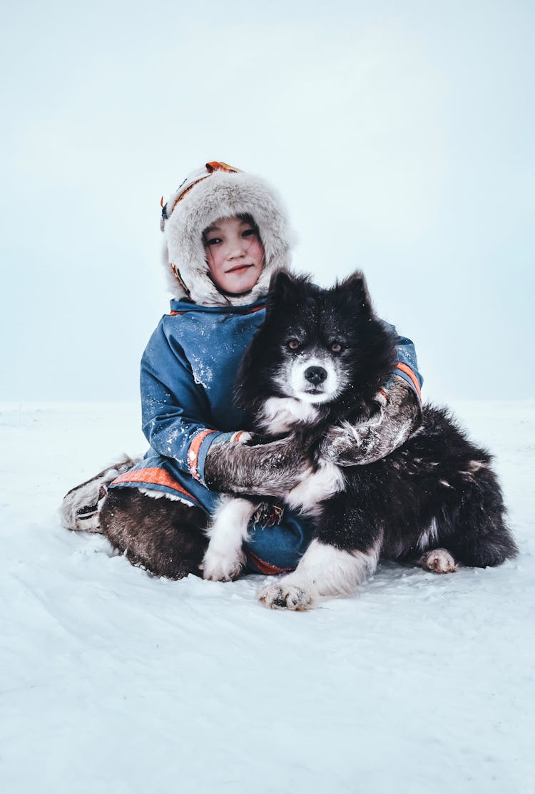 Girl In Winter Coat Sitting In Snow Hugging Dog