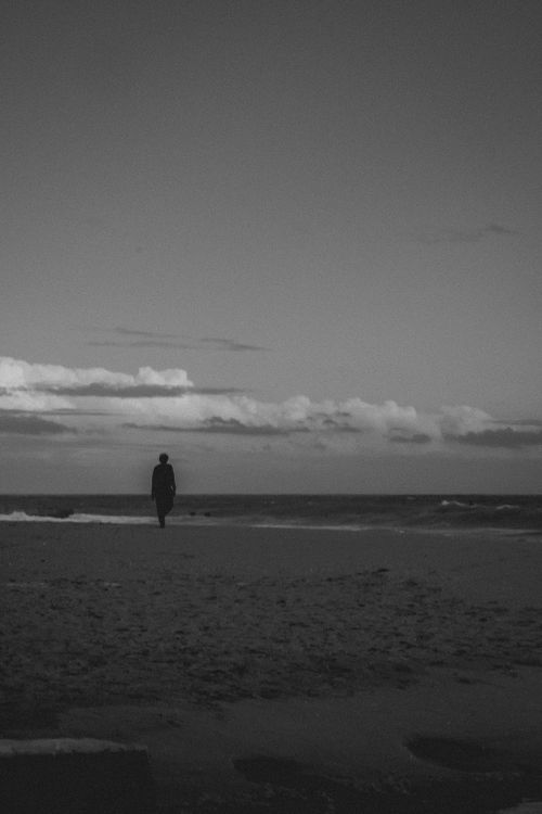 Free A serene black and white silhouette of a man walking along a beach under a vast sky. Stock Photo