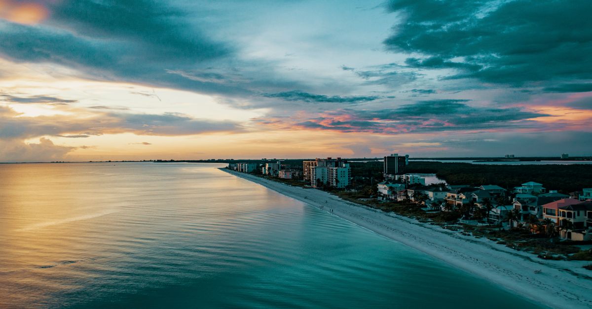 A breathtaking view of a coastal beach at sunset with buildings lining the shore, capturing the calm ocean and dramatic sky.
