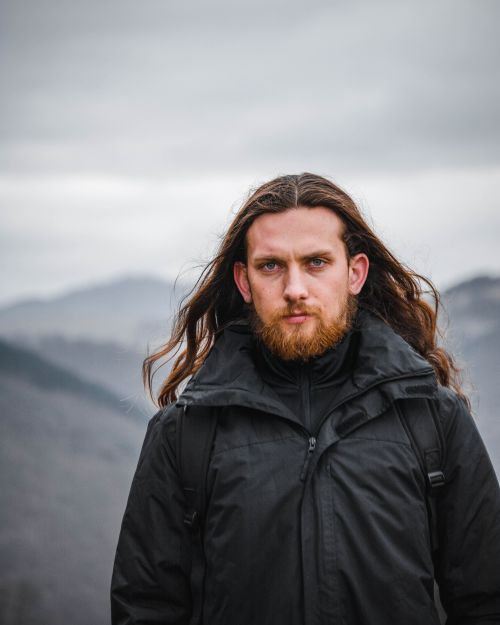 Free Portrait of a bearded man with long hair in outdoor mountain setting, wearing a black jacket. Stock Photo
