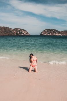Adult woman in bikini kneeling on a sandy beach with clear ocean views and rocky islands.