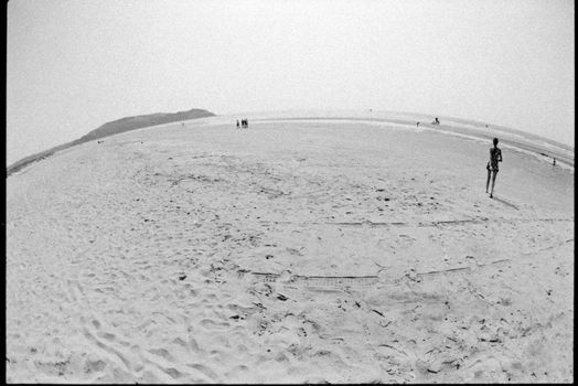 Black and white beach scene with fisheye effect, showing people walking on a sandy shore.