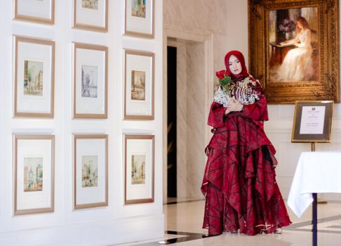 Woman in a red gown holding flowers in an art gallery with framed paintings. - Fotografer Sidoarjo