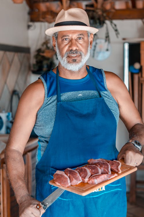 Man in Blue Kitchen Apron Holding Knife and Chopping Board With Sliced Meat