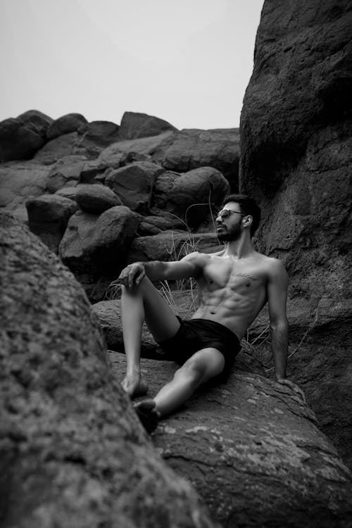 Grayscale Photo of a Topless Man Sitting on the Rock