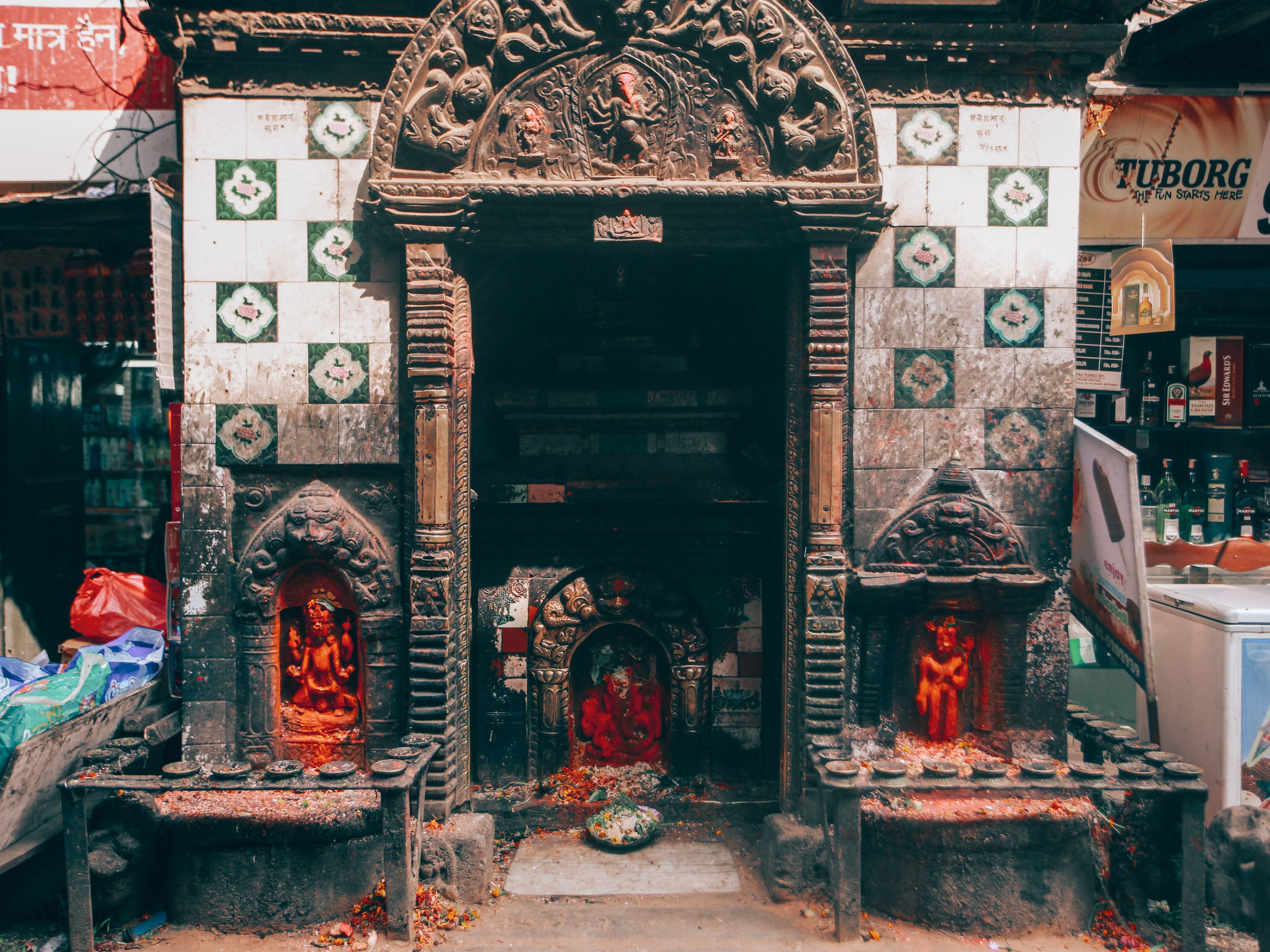A small street shrine with detailed carvings and offerings in an urban setting.