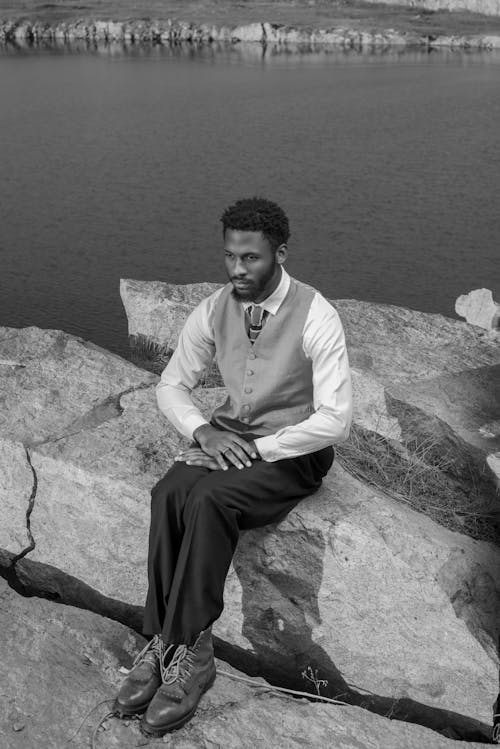 Black and White Photo of a Man Sitting on a Rock by the Lake