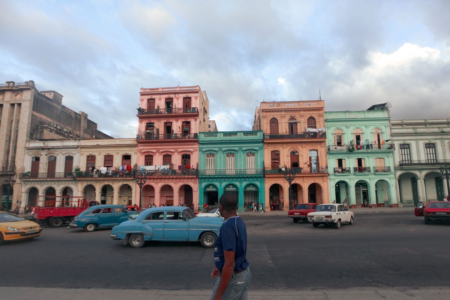 Een drukke straat in Havana met kleurrijke koloniale gebouwen en klassieke auto's onder een bewolkte lucht.