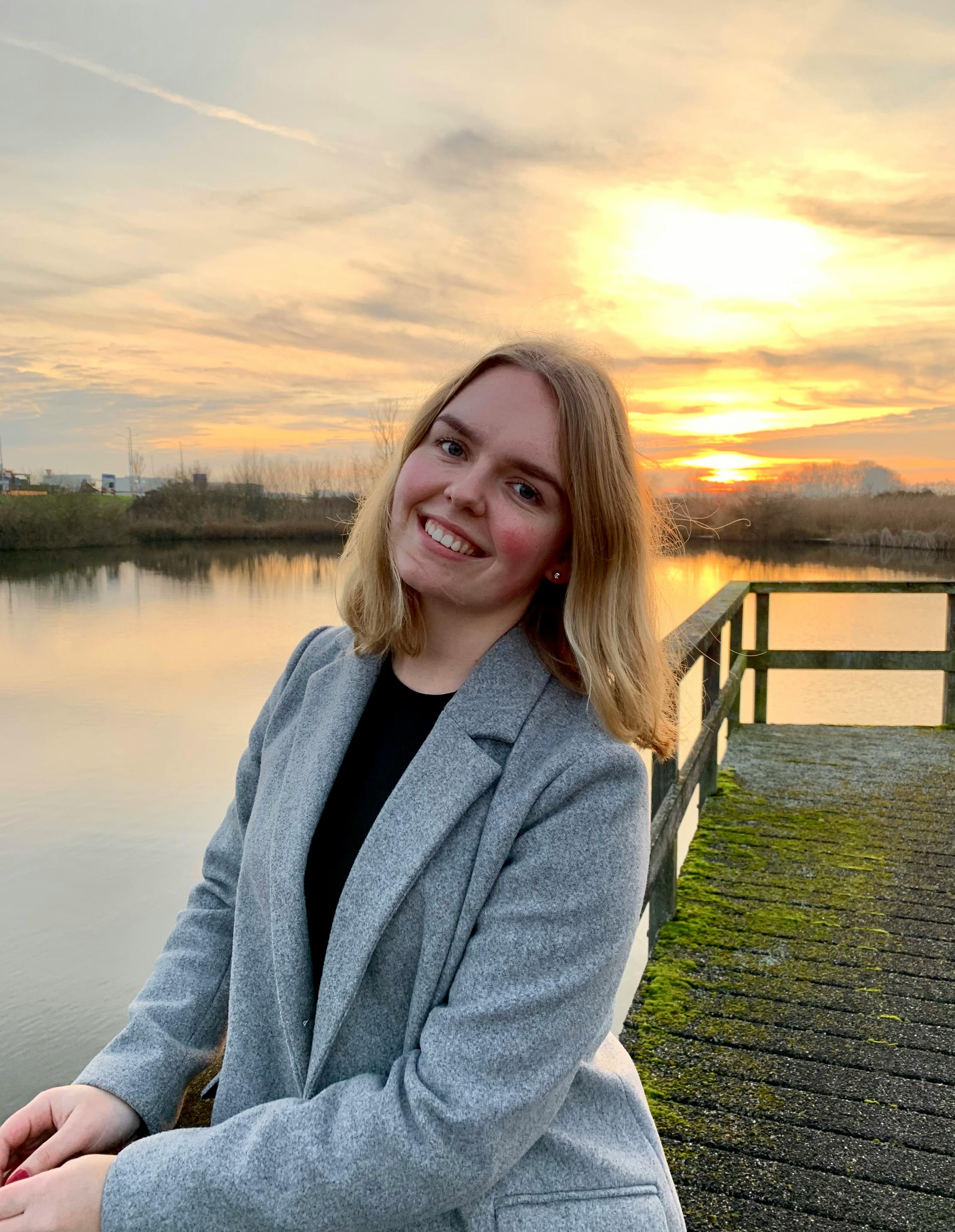 A woman in a blazer smiling at the camera during a beautiful sunset by a serene lake.