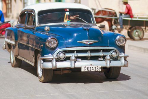 Free Classic blue Chevrolet Deluxe car driving on a sunny street with a horse-drawn cart in the background. Stock Photo