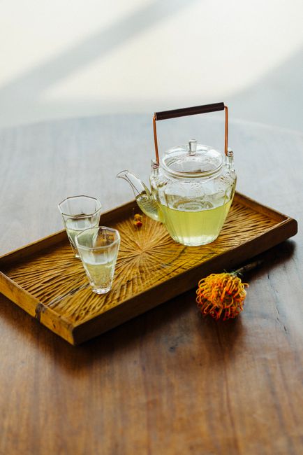 Glass teapot with green tea and cups on a wooden tray, adorned with a chrysanthemum, in natural light.