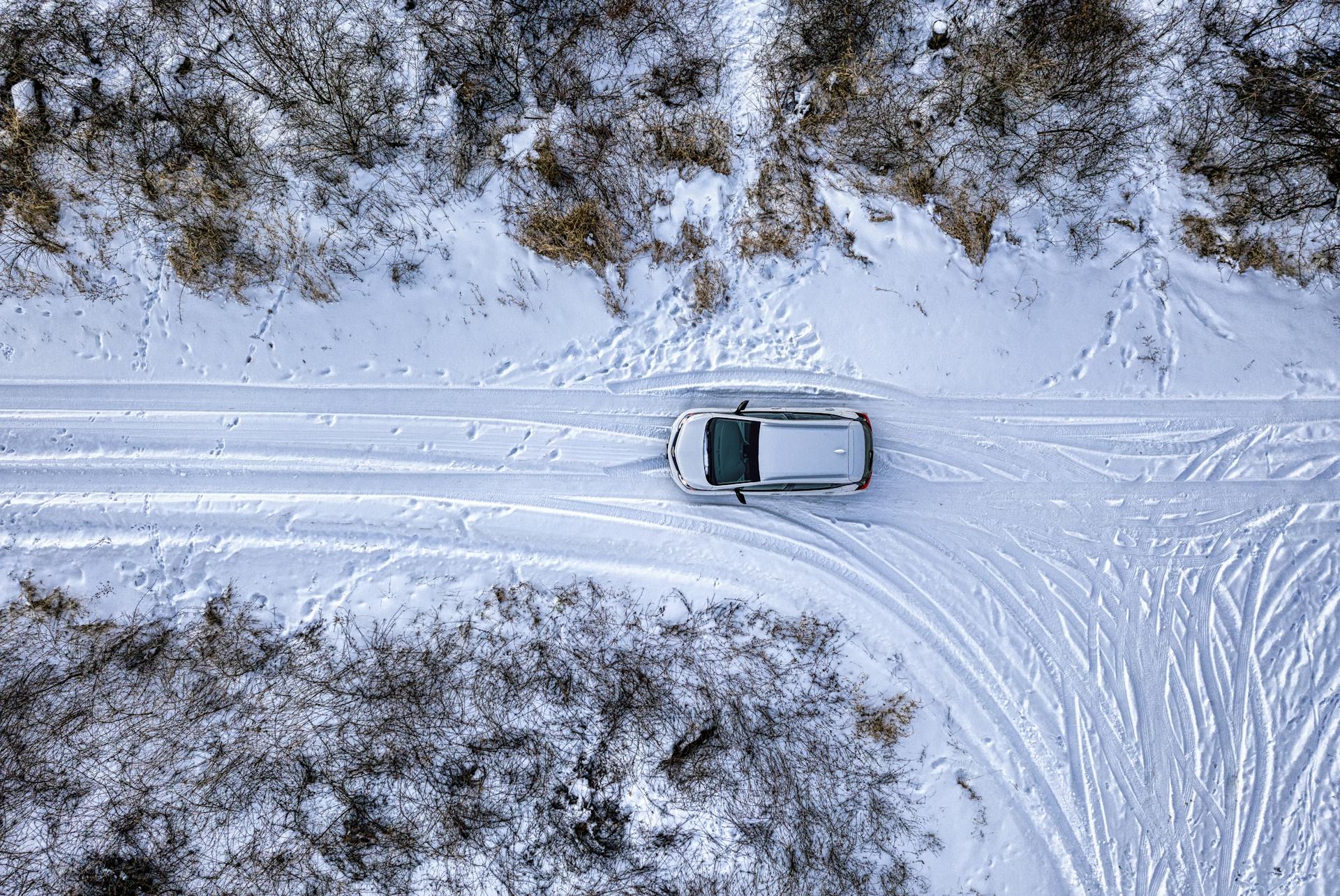 Drone shot of a car driving on a snow-covered road in winter, surrounded by trees.