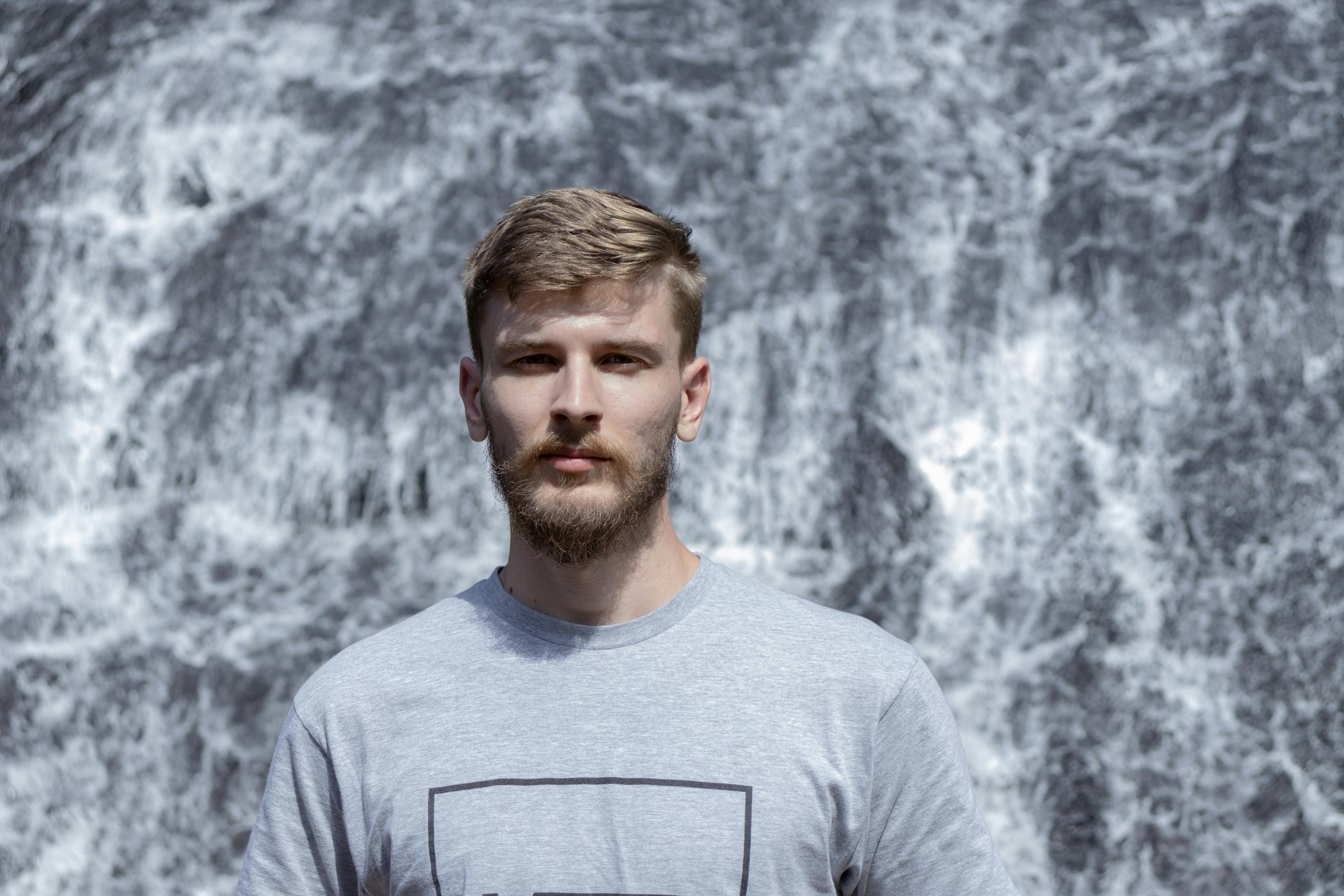 A young bearded man stands confidently in front of a cascading waterfall wearing a gray shirt.