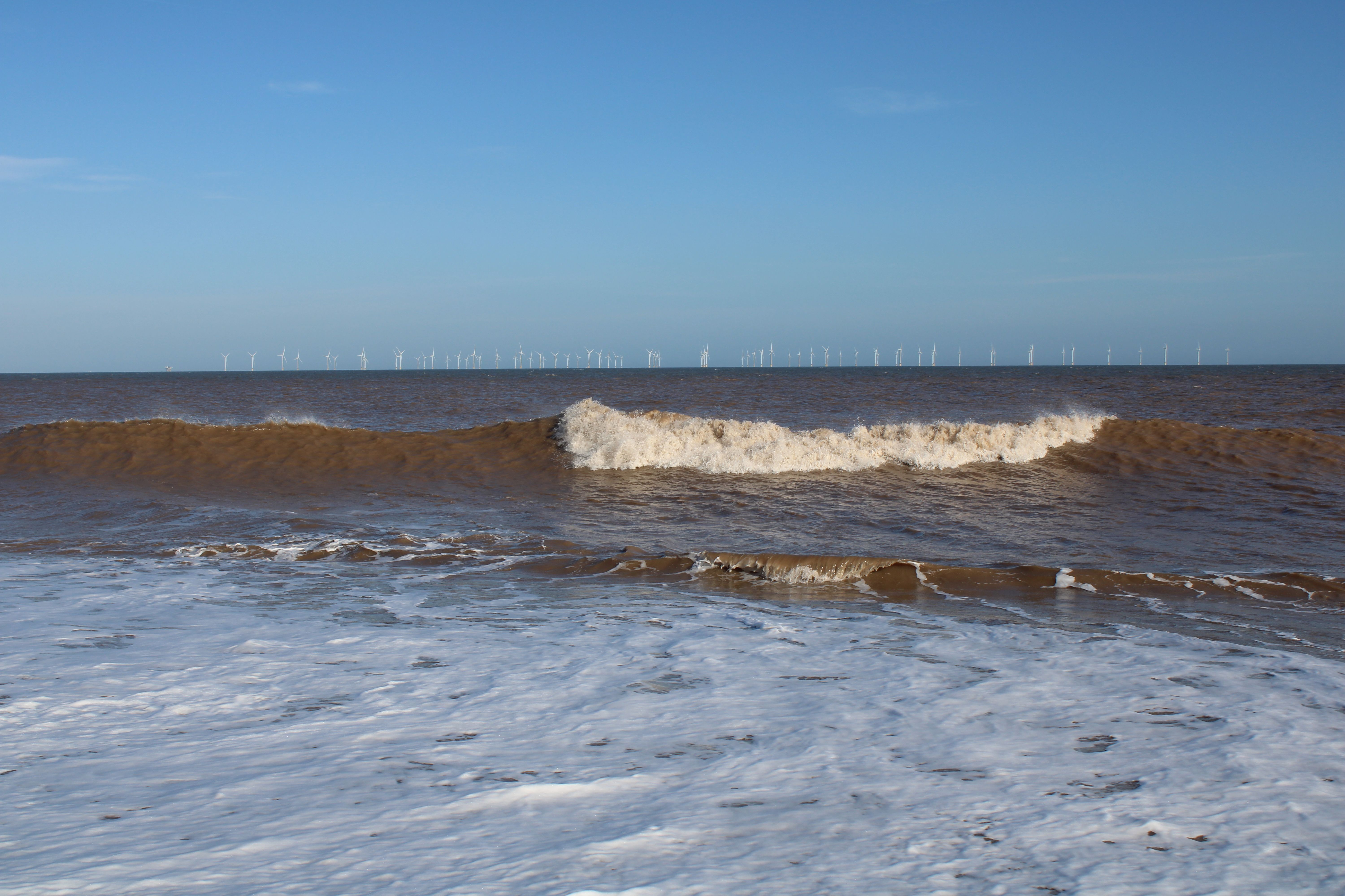 Ocean waves with whitecaps under a bright sky and small turbines on the horizon.
