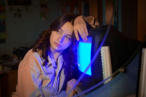 Free A woman sits indoors, gazing thoughtfully at a vintage television emitting a blue glow. Stock Photo