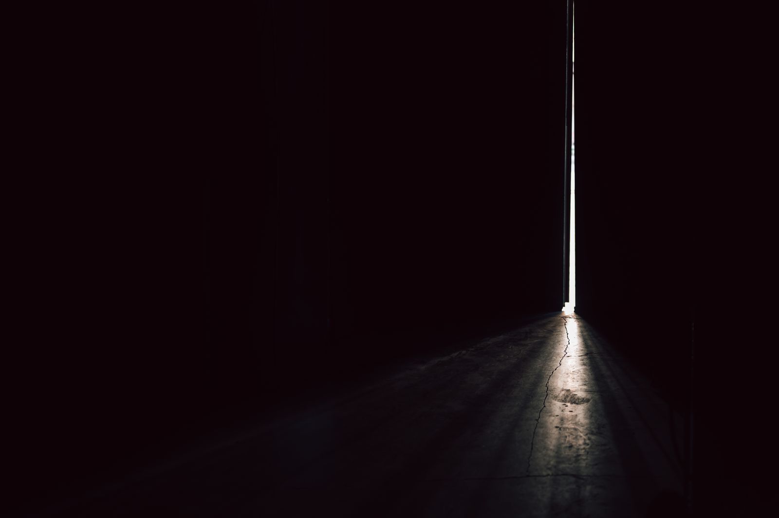 A narrow beam of light cutting through a dark room from a barely-open door, illuminating a cracked concrete floor.