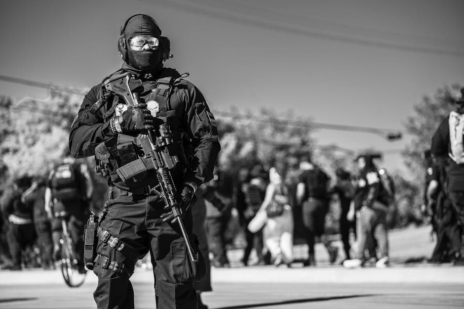 Monochrome image of a soldier in uniform with a rifle standing on a street amidst a crowd, conveying military presence.