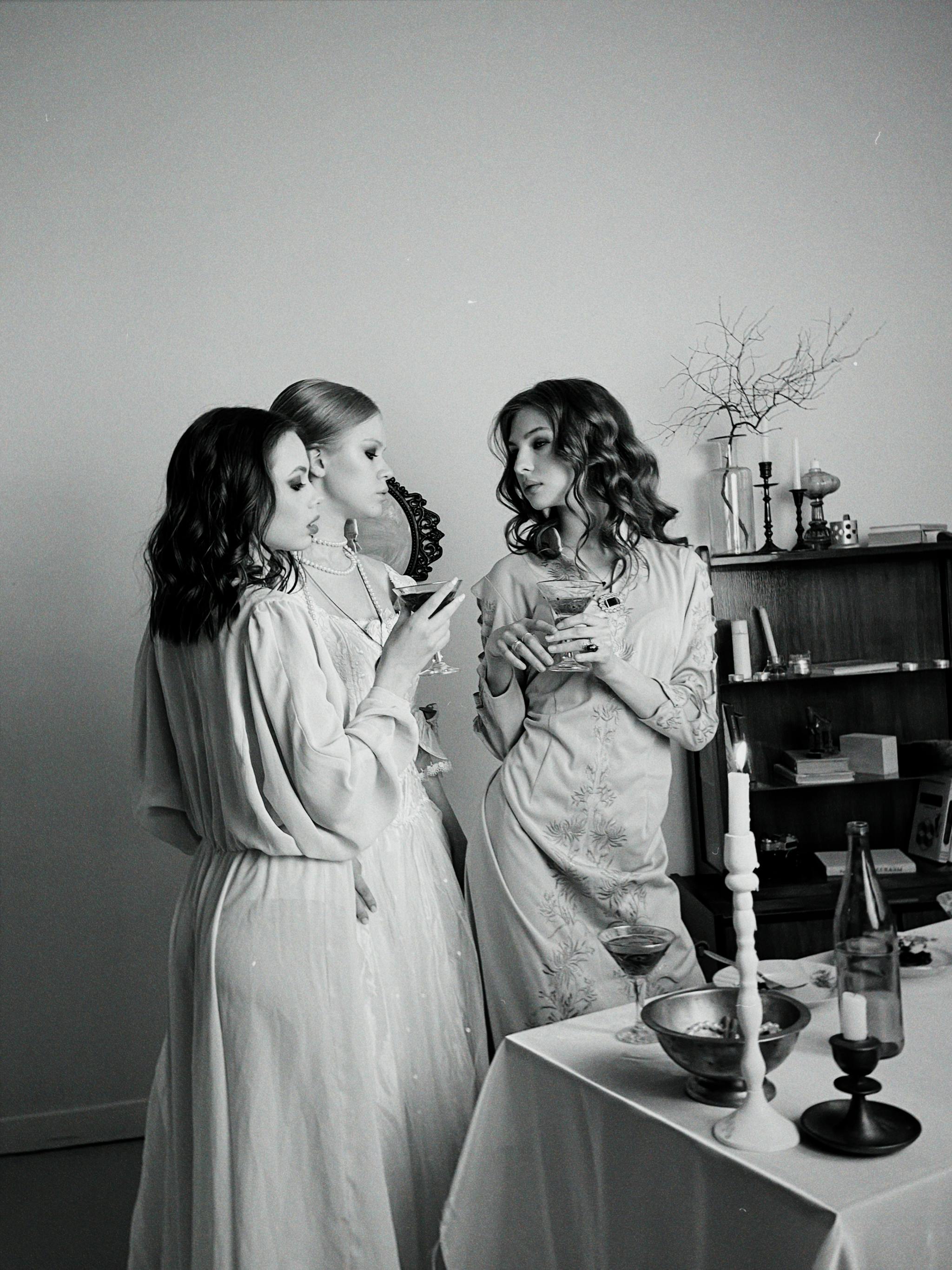 Three women in vintage dresses enjoying an elegant evening socializing and sipping wine.