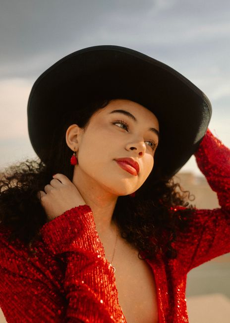 Elegant portrait of a woman in a red sequin outfit and black hat against a neutral background.