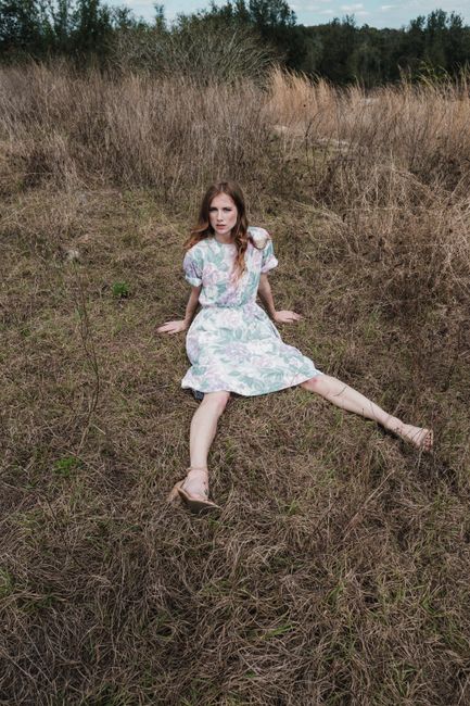 A young woman in a floral dress sits in a dry field under a cloudy sky.