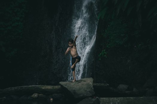 A child playfully leaps on a rock in front of a lush waterfall in West Java, Indonesia.