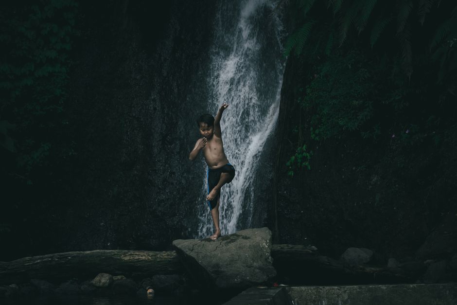 A child playfully leaps on a rock in front of a lush waterfall in West Java, Indonesia.