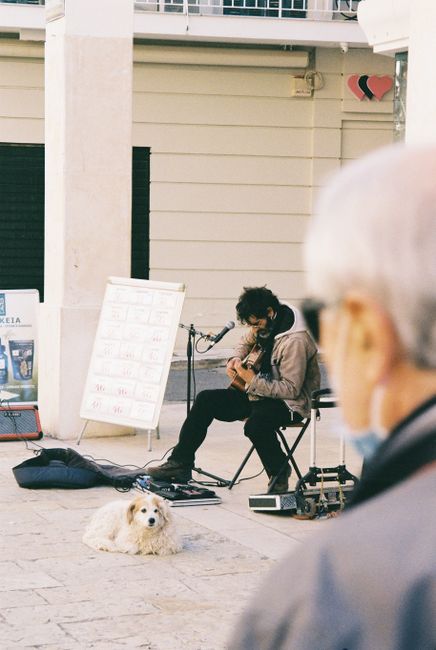 A street musician plays guitar with his dog nearby on a sunny day in Corfu, Greece.