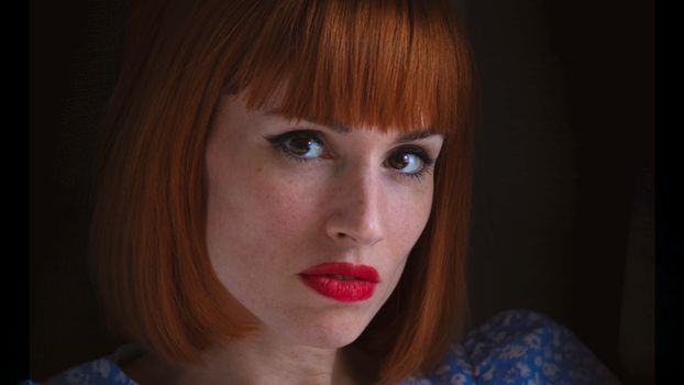 Close-up portrait of a woman with striking red hair and bold red lipstick against a dark background.