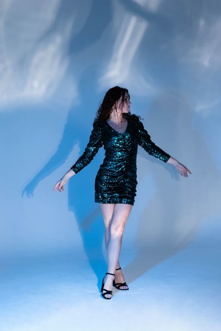 Woman in a shimmering sequin dress posing against a dramatic backdrop of shadows under blue lighting.