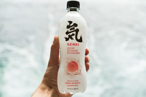 Free Hand holding a Genki peach-flavored sparkling water bottle against a blurred background. Stock Photo