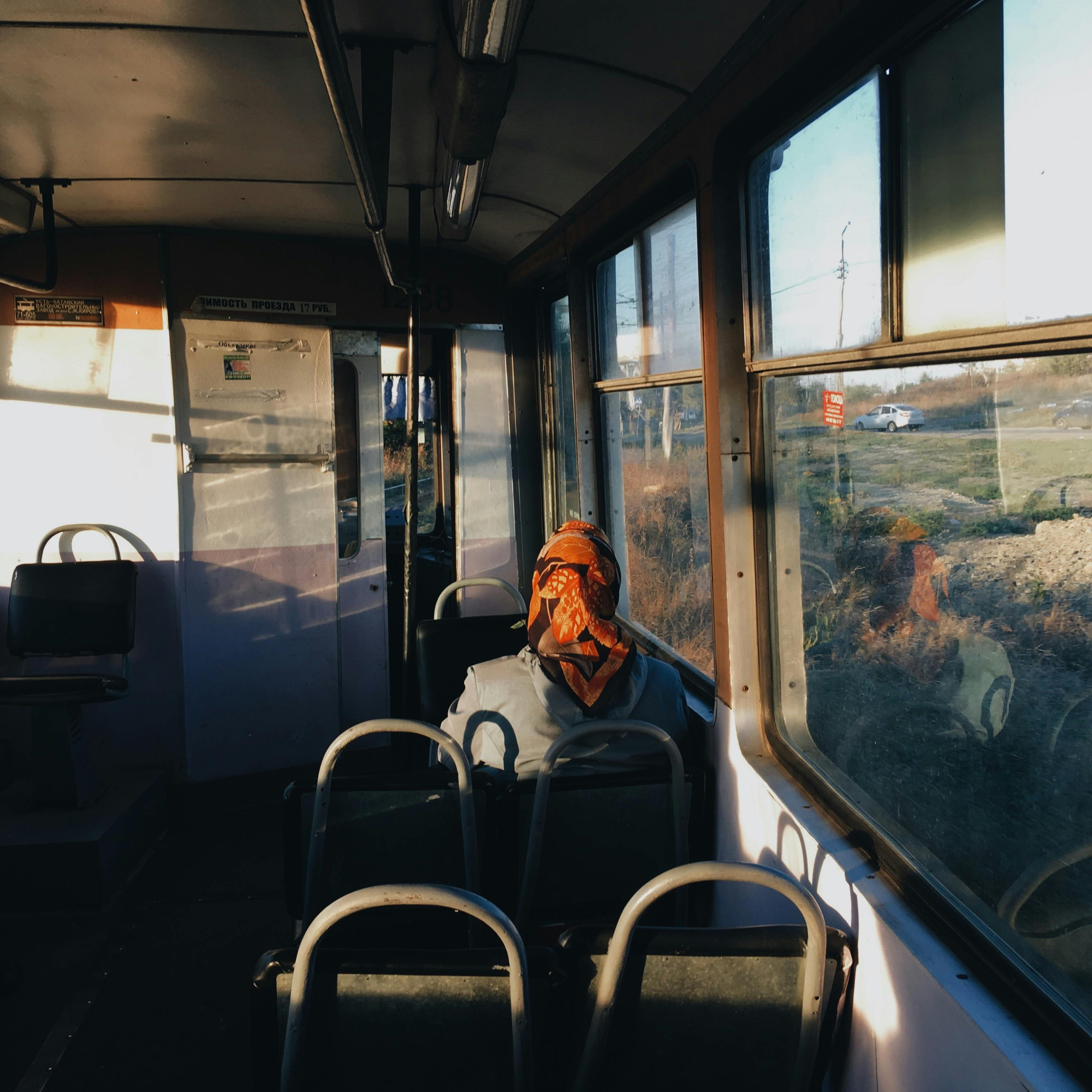 A woman wearing a headscarf sits alone in a vintage bus, gazing out the window at sunset.