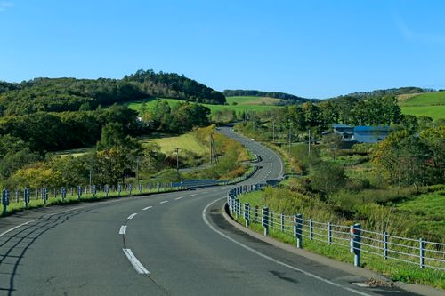 Free A beautiful rural road winding through lush green hills under a clear blue sky. Stock Photo