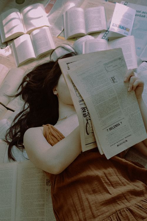 Free Young woman comfortably reads a newspaper amidst scattered books, creating a cozy, intellectual vibe. Stock Photo