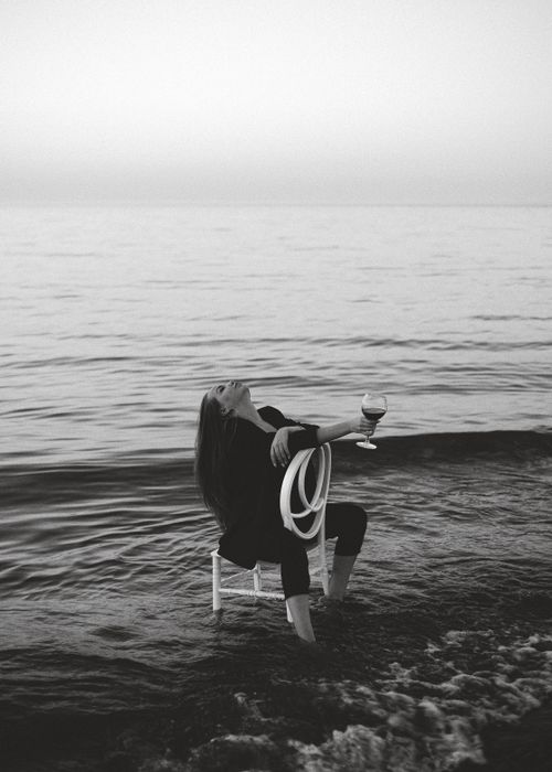 Free A relaxed woman enjoys a glass of wine by the seaside during twilight, sitting on a chair in the water. Stock Photo