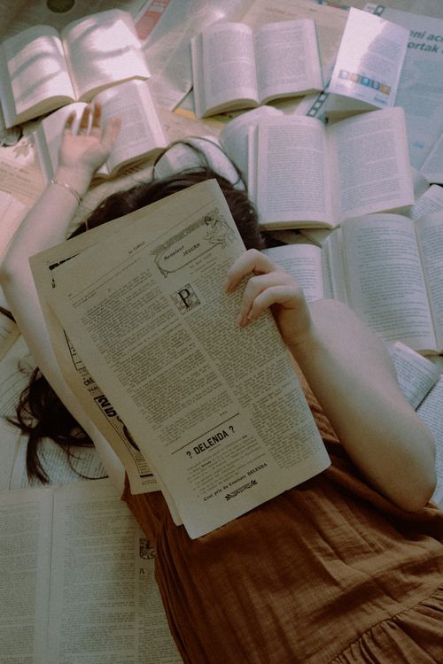 Free An adult woman lies on the floor surrounded by open books, reading a newspaper in a cozy indoor setting. Stock Photo