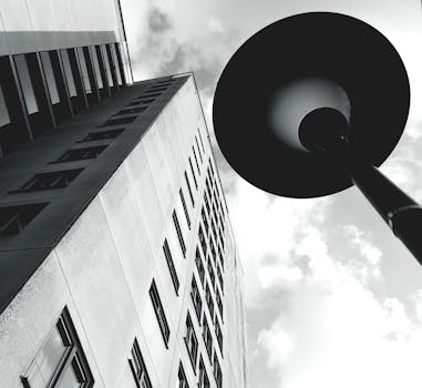 这张引人注目的低角度视图以灰度呈现了现代摩天大楼和多云的天空，画面中还有路灯